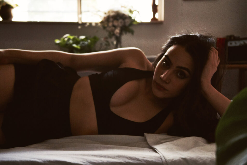 Valeria Cisneros lounges gracefully on a bed wearing a black outfit with a serene expression surrounded by indoor plants and a window letting in soft light.